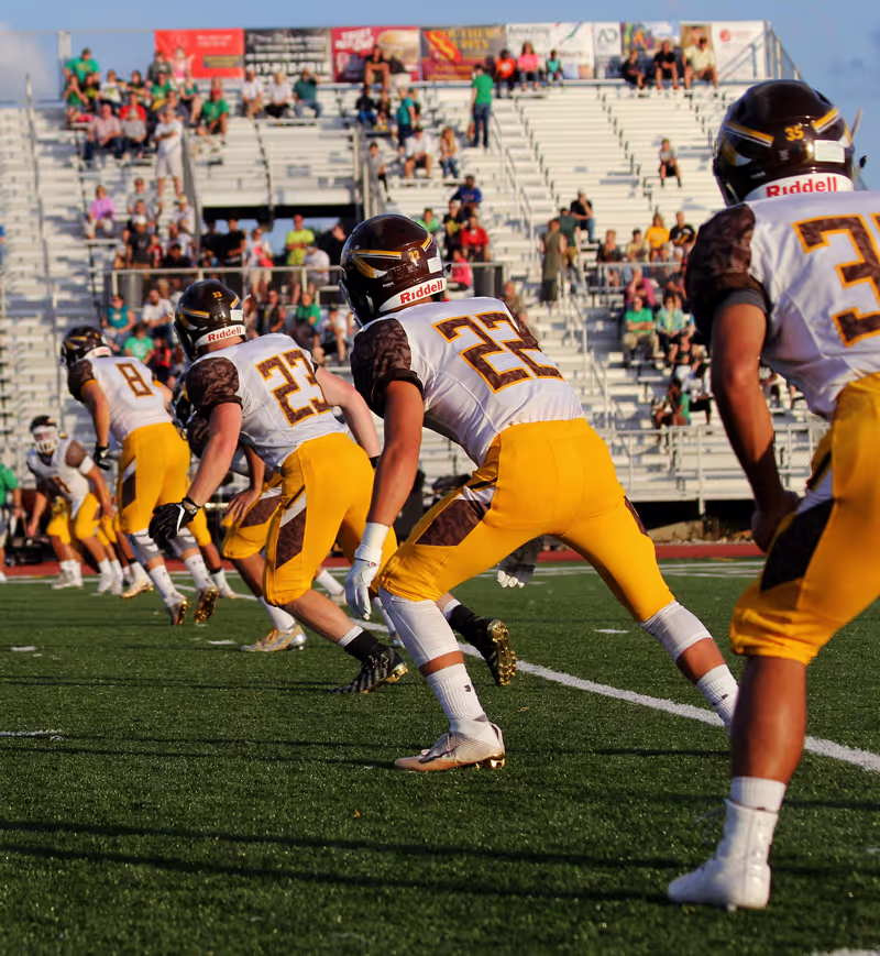 Football players in yellow pants and white jerseys lined up on a green field in front of a crowd.