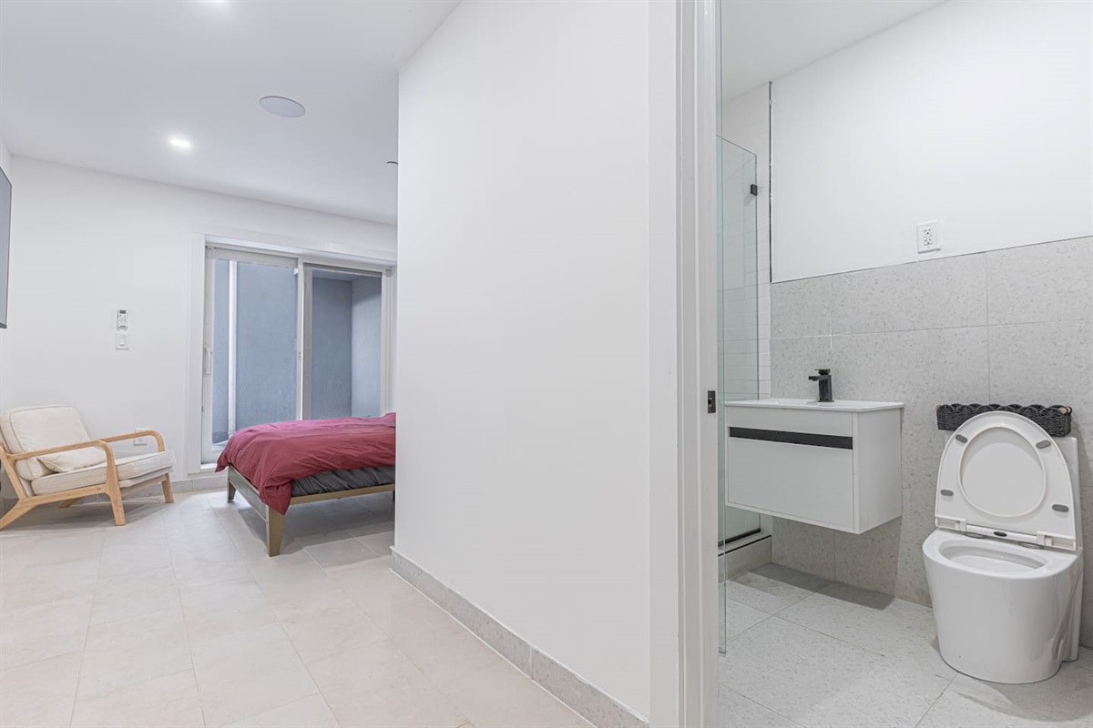 Bright bedroom with a red duvet, armchair, and sliding patio door; adjacent clean white bathroom featuring a minimalist