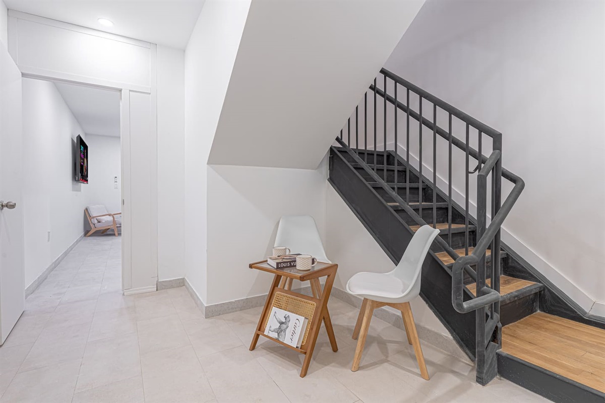 Modern home interior featuring a white staircase with a black railing, white chairs by a wooden side table, and a hallwa