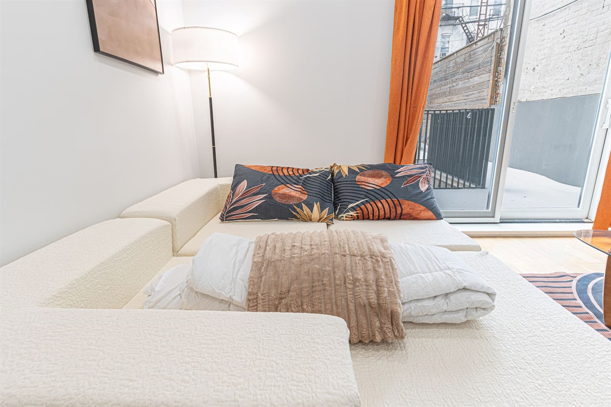 White sectional sofa with floral pillows and a tan throw blanket, positioned near a glass door with orange curtains.