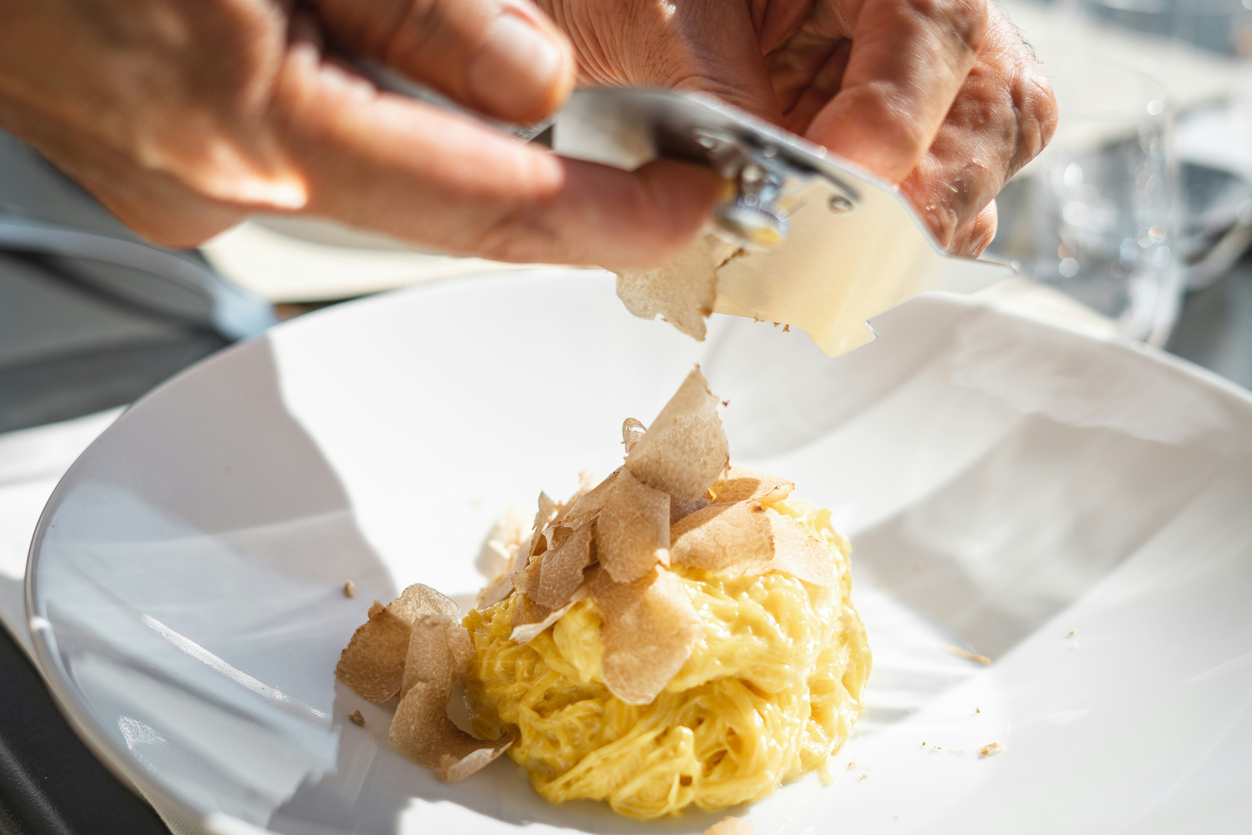 Nahaufnahme von Händen, die braune Trüffelscheiben mit einer Reibe über einer Portion cremiger Pasta in einem weißen Teller hobeln.