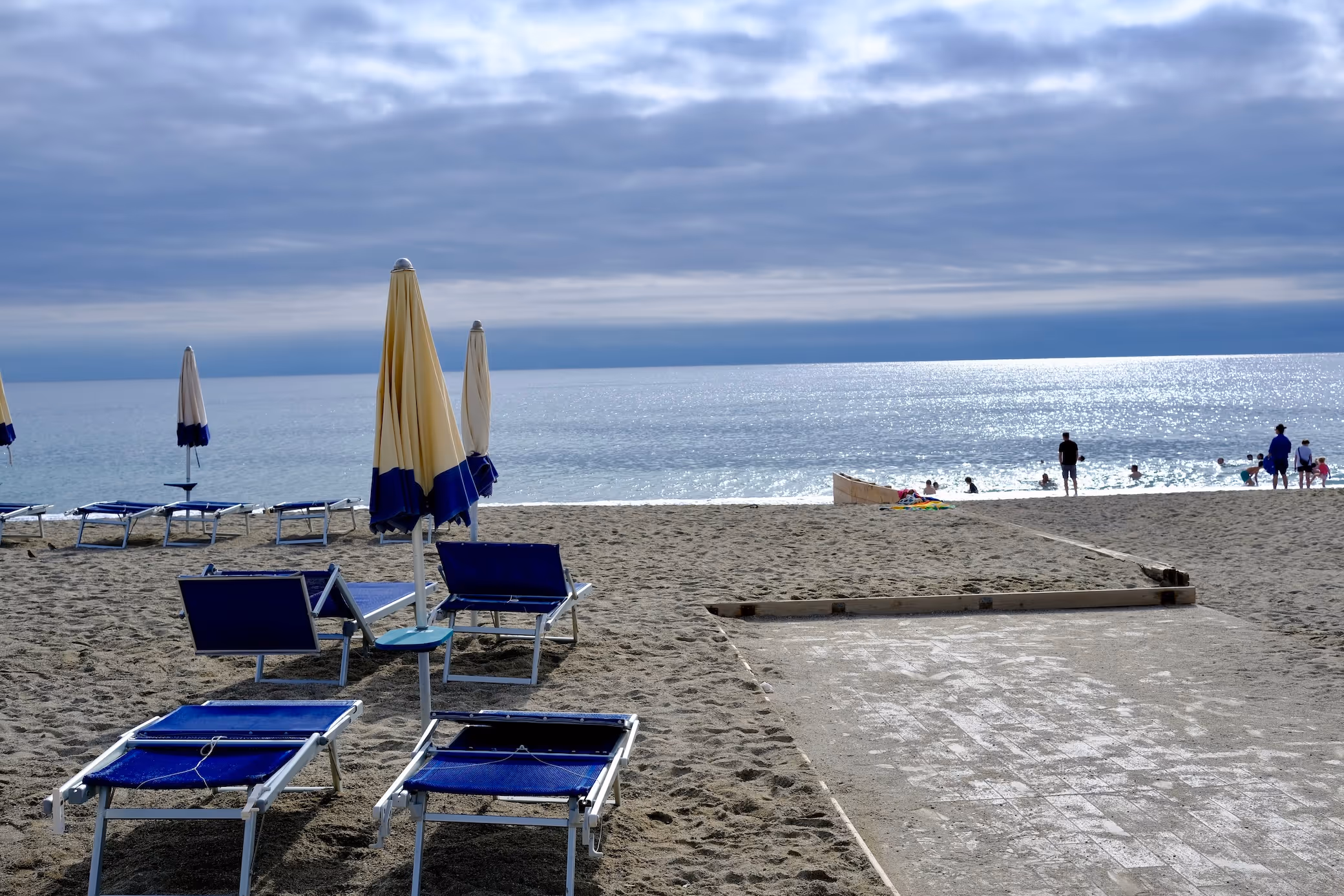 Ein Strand mit Liegestühlen und geschlossenen Sonnenschirmen in Blau und Gelb auf Sand, im Hintergrund das ruhige Meer und bewölkter Himmel, einige Menschen sind am Wasserrand.