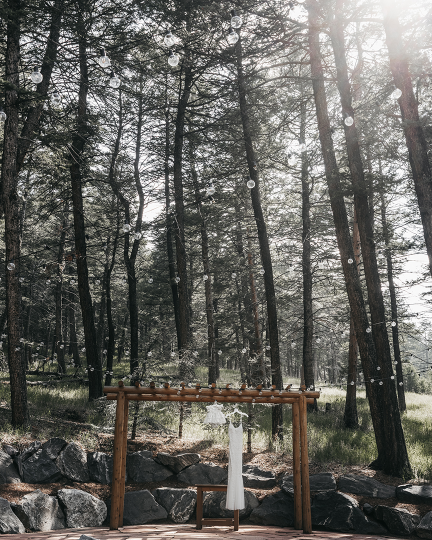 A bridal dress with sequins and a baby's dress hanging amongst the pines of Colorado off a wooden archway 