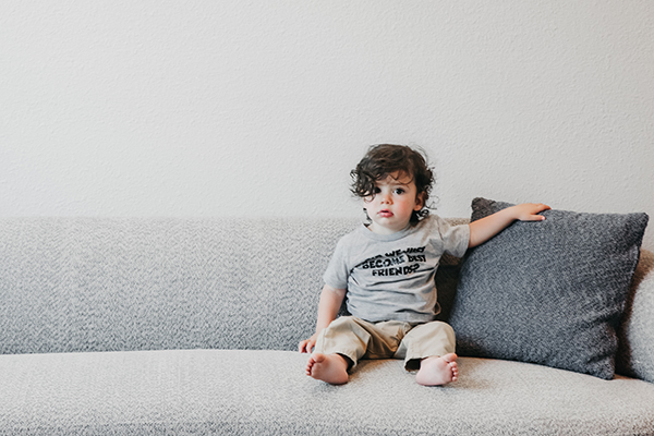 A cute little 2 year old boy sitting adorable on a big white couch.