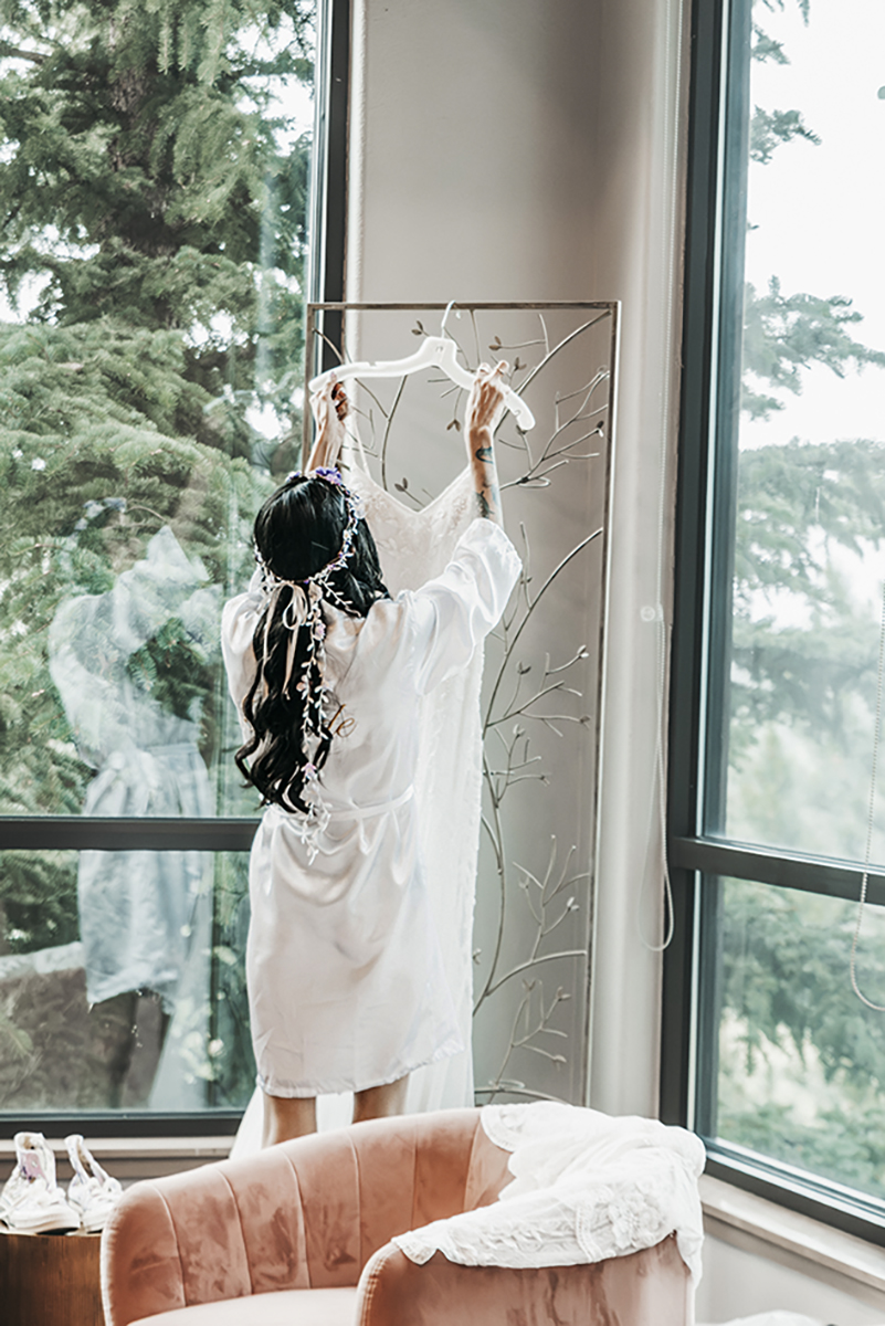 A bride and her wedding dress in an imag with pine trees peaking into the floor length photos of her dressing room