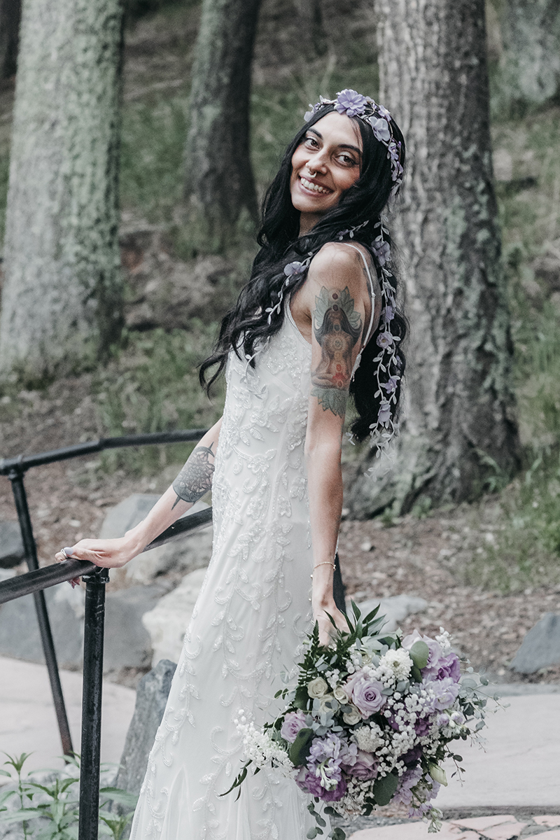 A bridal portrait with a smiling bride and her lavender and sage flowered veil and white sequinned gown with spaghetti straps and ankle length holding a large beautiful bouquet with like colors against thick Pine trunks.