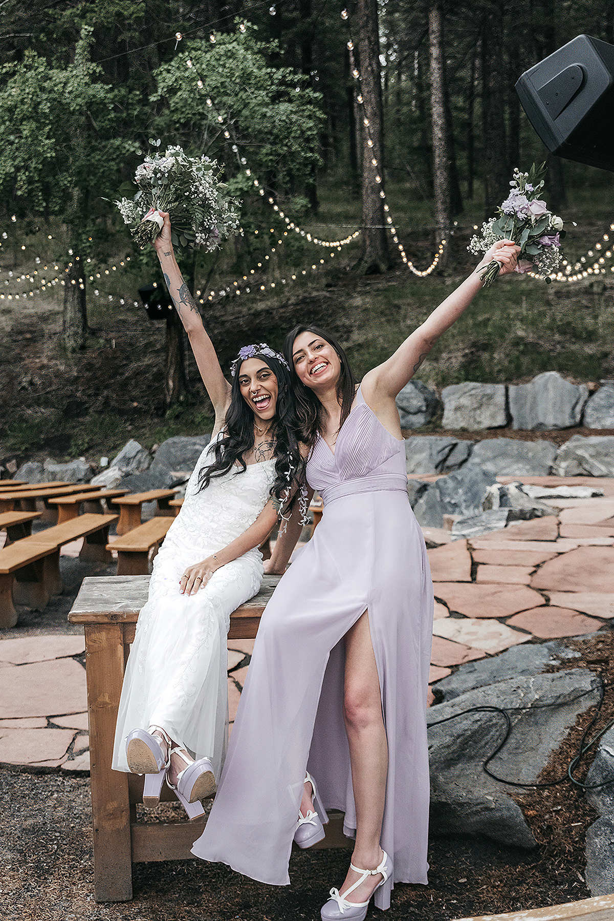 A wonderful cheery moment of two sisters raising their bouquets with sage and lavender accents with Pine trees and Colorado rocks in the mountain side. 