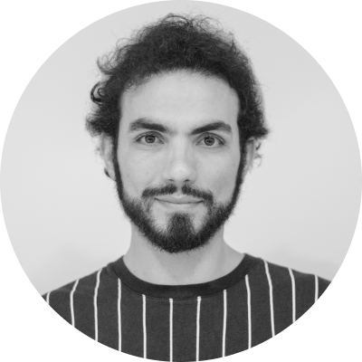 Portrait of a man with curly hair, beard, and mustache wearing a striped shirt, smiling slightly against a plain background.