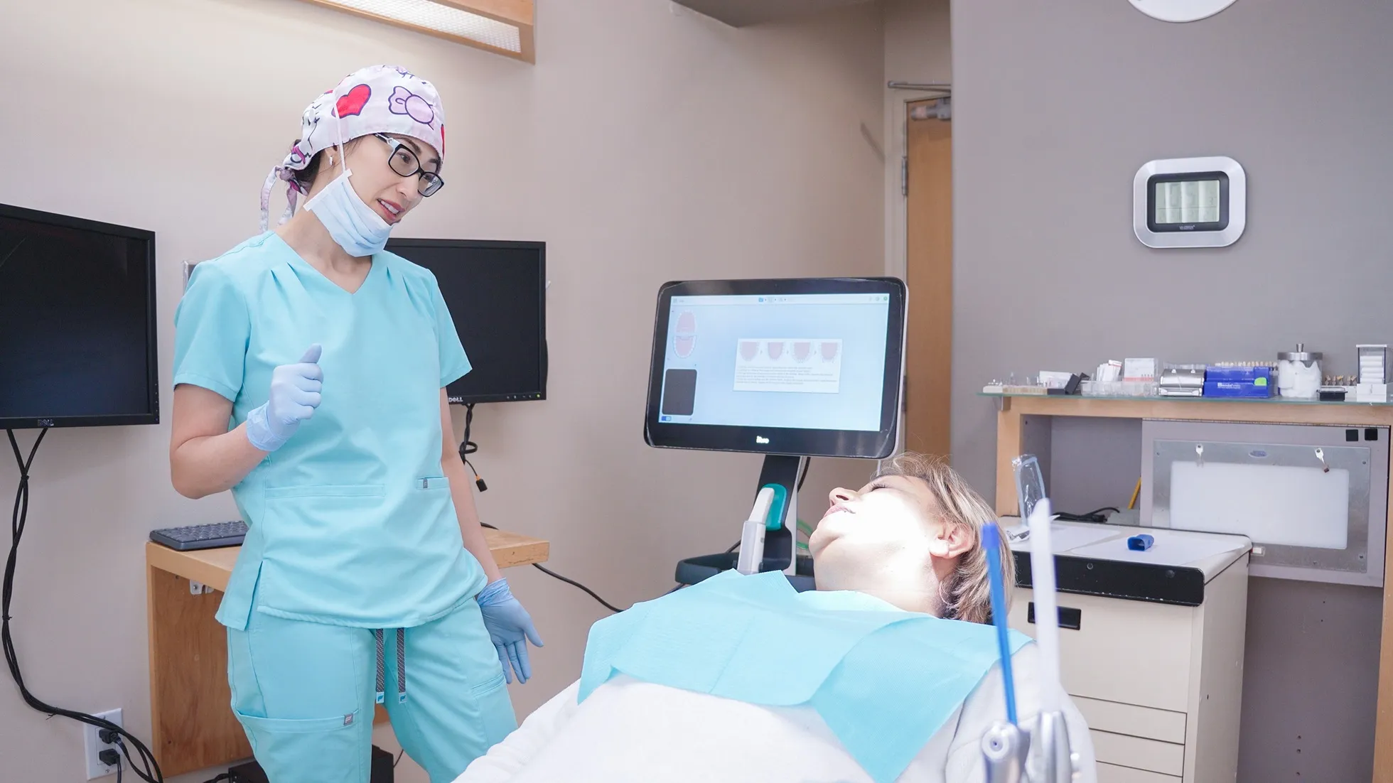 Dental professional in full PPE, including a face shield and mask, attending to a patient lying in a dental chair. Medical monitors and equipment are visible in the background, indicating a modern clinical setting