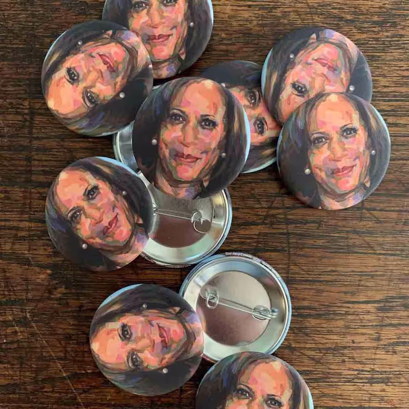 Several small pins with Kamala Harris's face on them lay randomly on a table, some face up, some face down.
