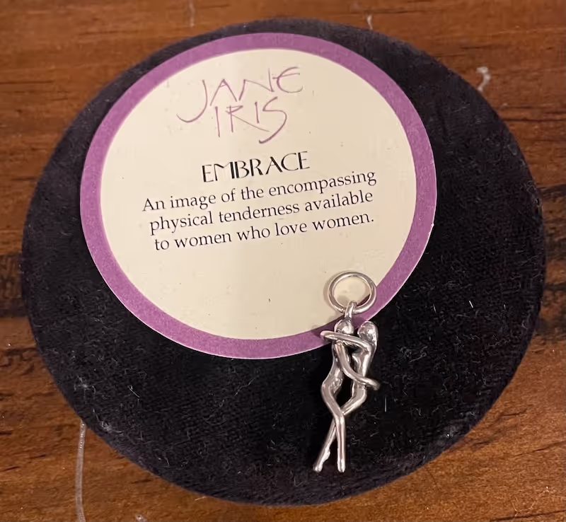 A silver pendant of two humans embracing is laid on a circular label describing it as Embrance by Jane Iris.