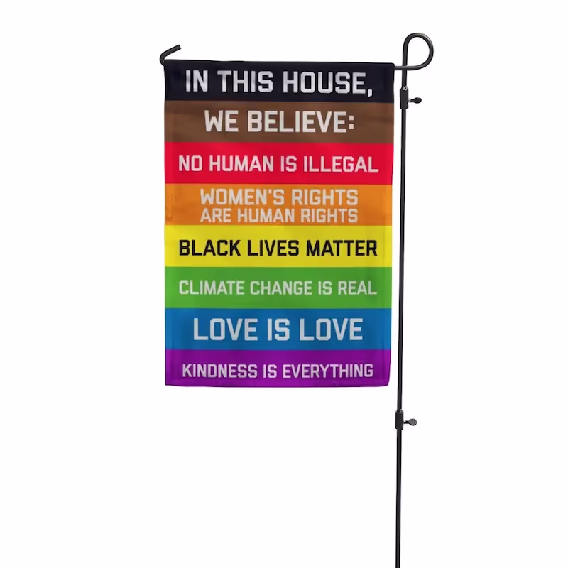 A muli-colored flag hangs from a rod. In This House Garden Flag has black text printed on the bars of color.