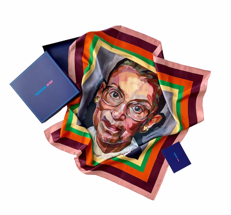 A printed scarf with the face of Ruth Bader Ginsberg (RBG) is positioned on a white surface with the purple packaging elements for the scarf in view at the edges.