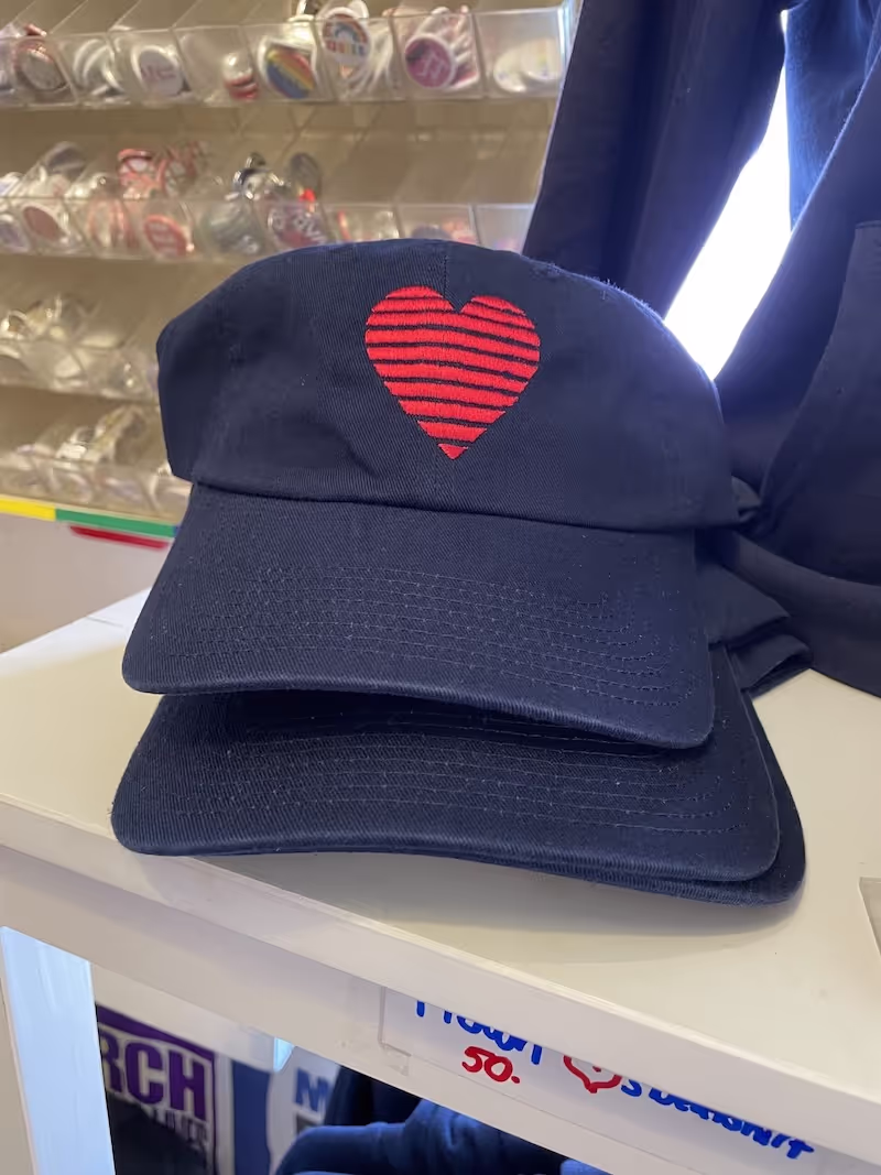 Two navy blue baseball hats are stacked. There is a horizontally stripes red Heart visible on the top one. The hats sit on a white shelf in a store.