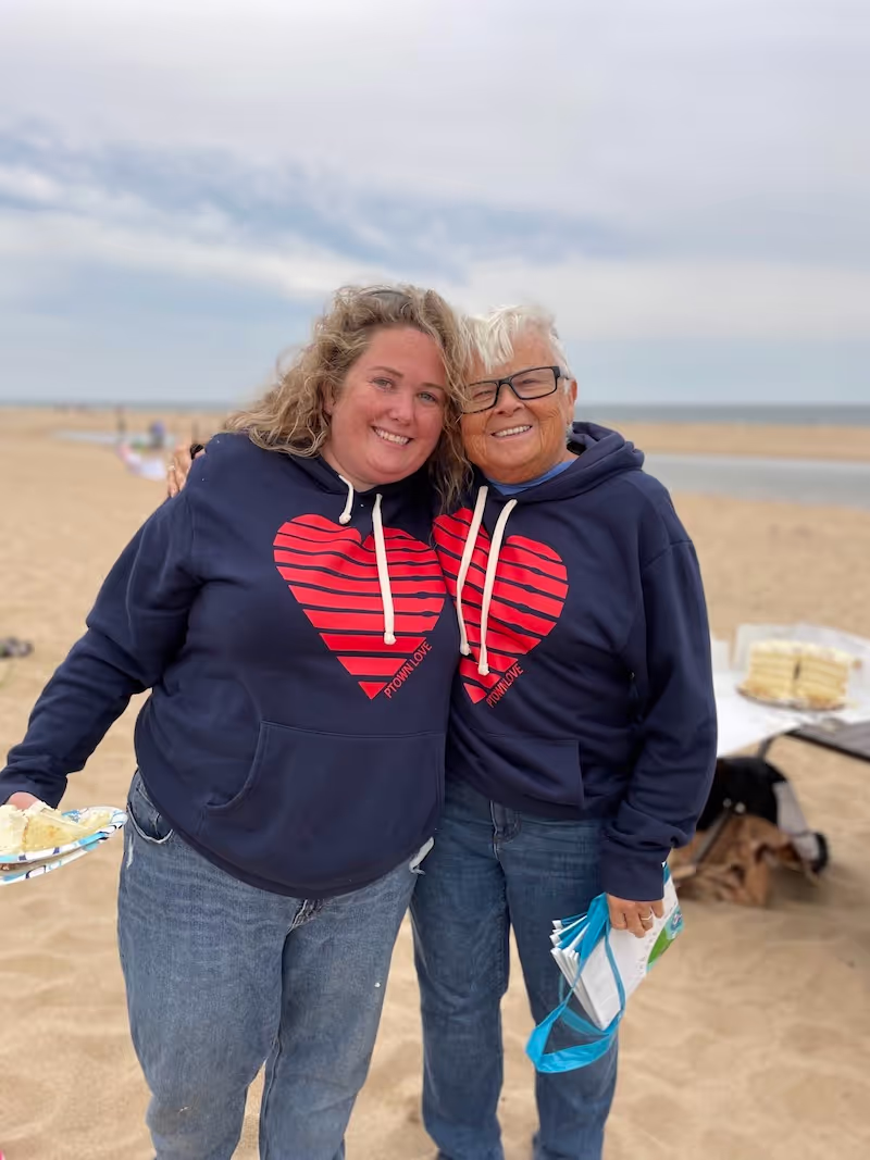 Two women wearing blue Provincetown Love hooded sweatshirts at a beach