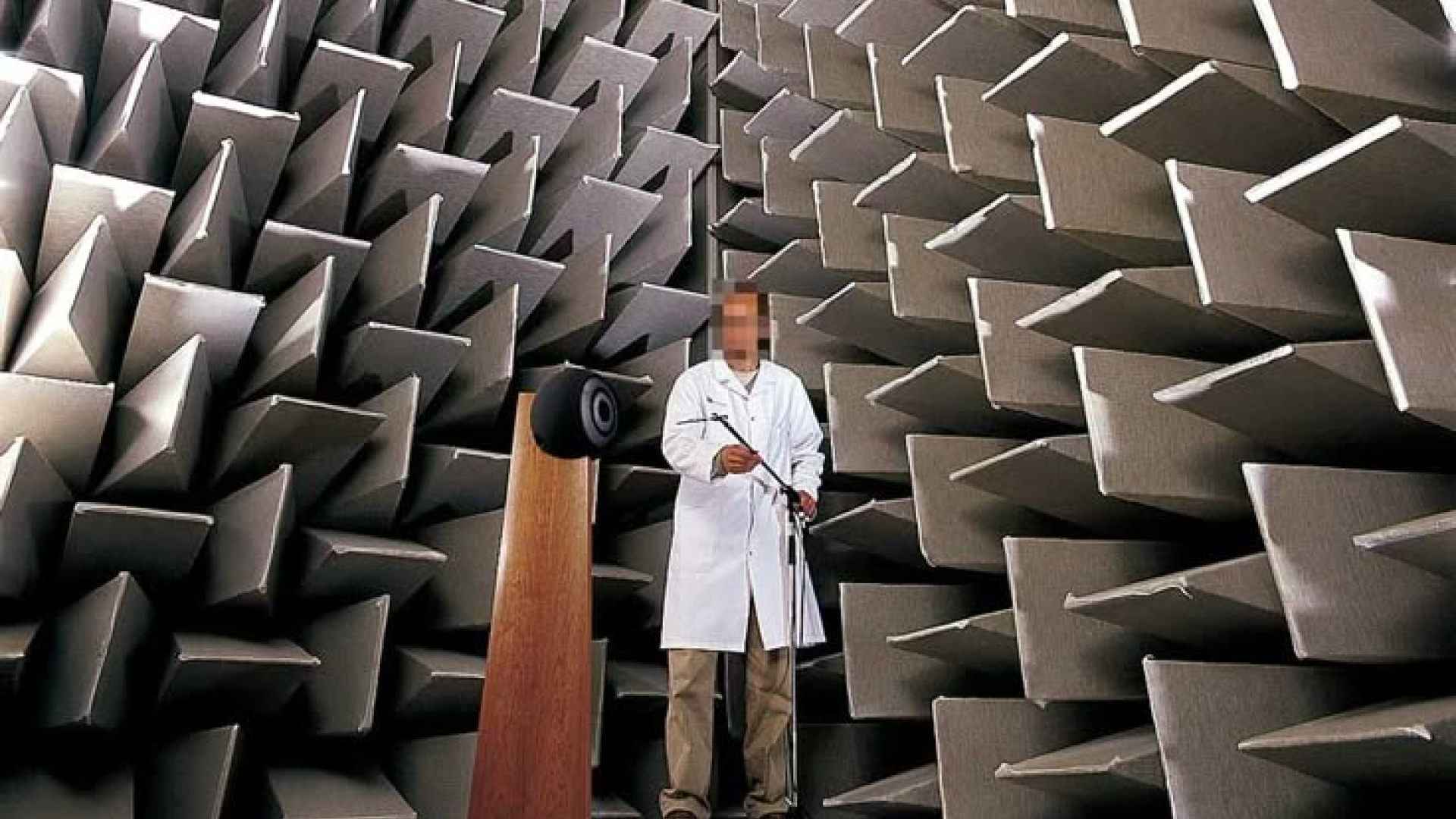 Man in lab coat standing next to a standing speaker in a room covered in felted baffles. 