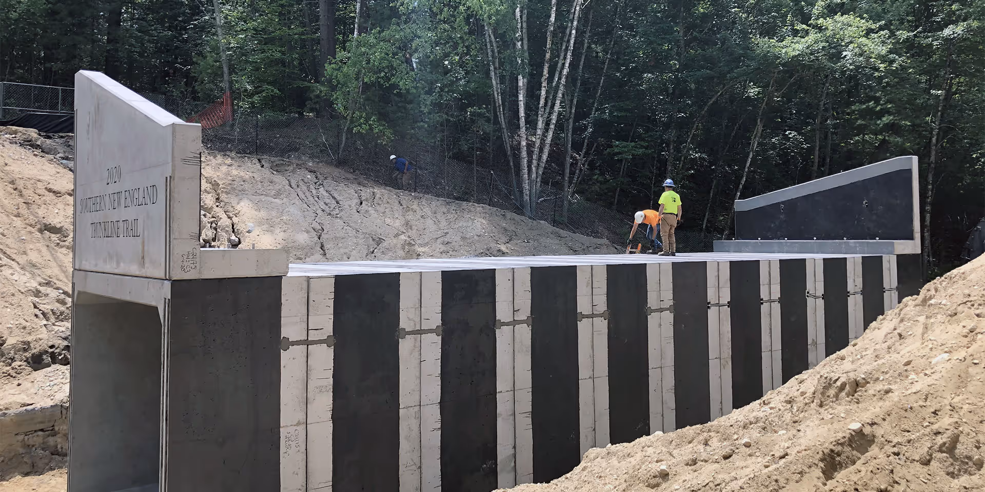 Two workers stand on top of a newly constructed concrete bridge surrounded by trees and dirt embankments.