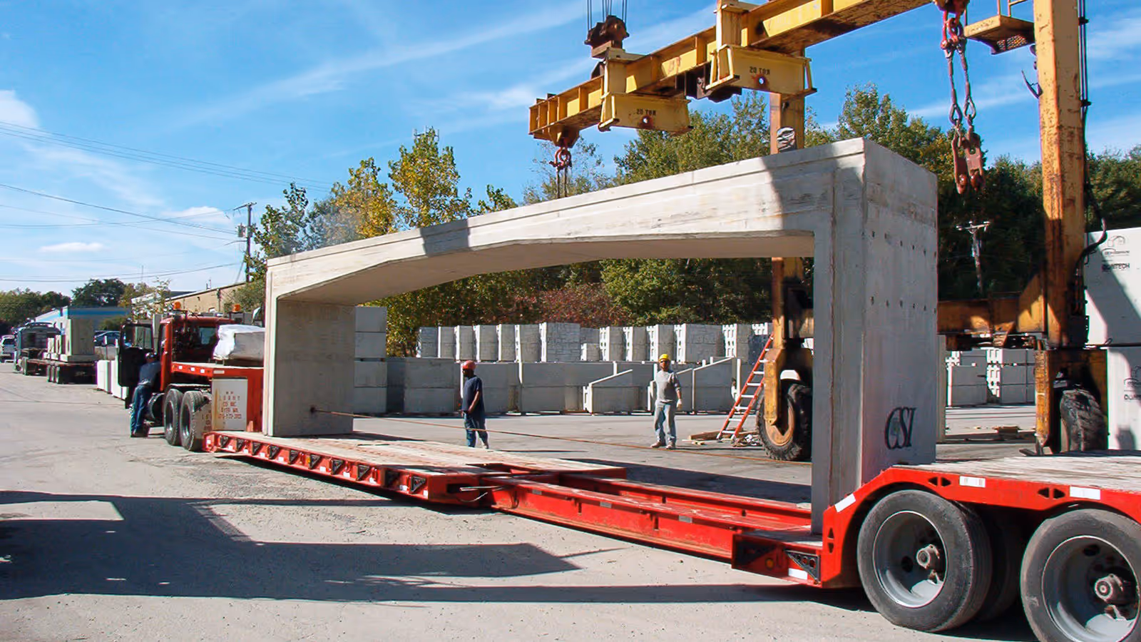 A large concrete bridge section is being loaded onto a red flatbed trailer by a yellow crane at an industrial site.
