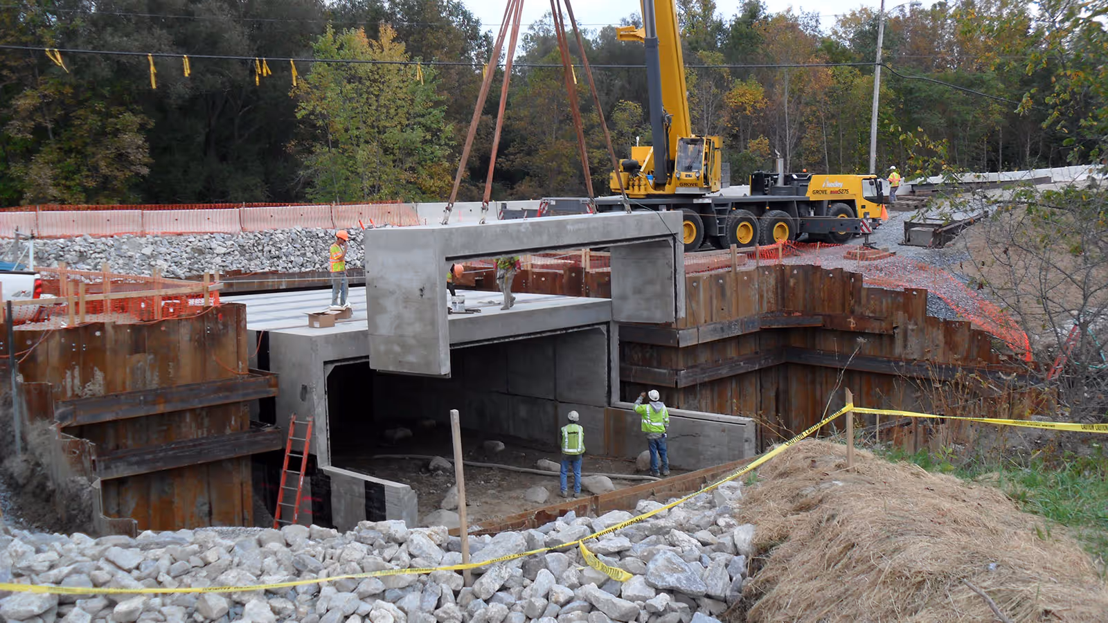 A crane lifts a large concrete section into place at a construction site with workers and safety barriers present.