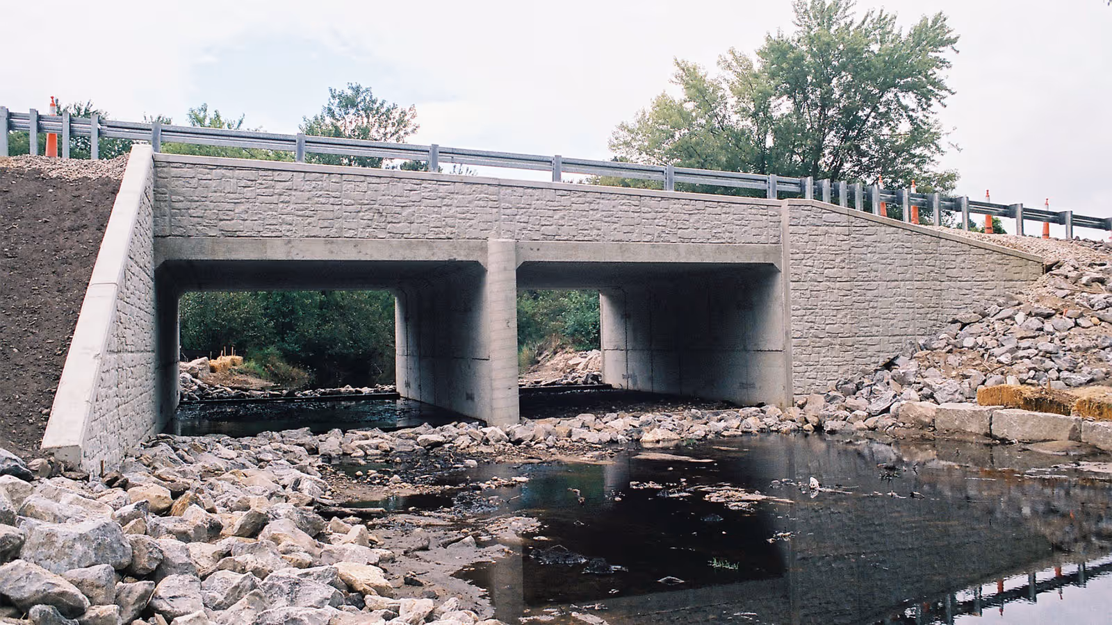 Concrete bridge with two rectangular spans crosses over a shallow rocky stream, surrounded by trees and stone embankments.