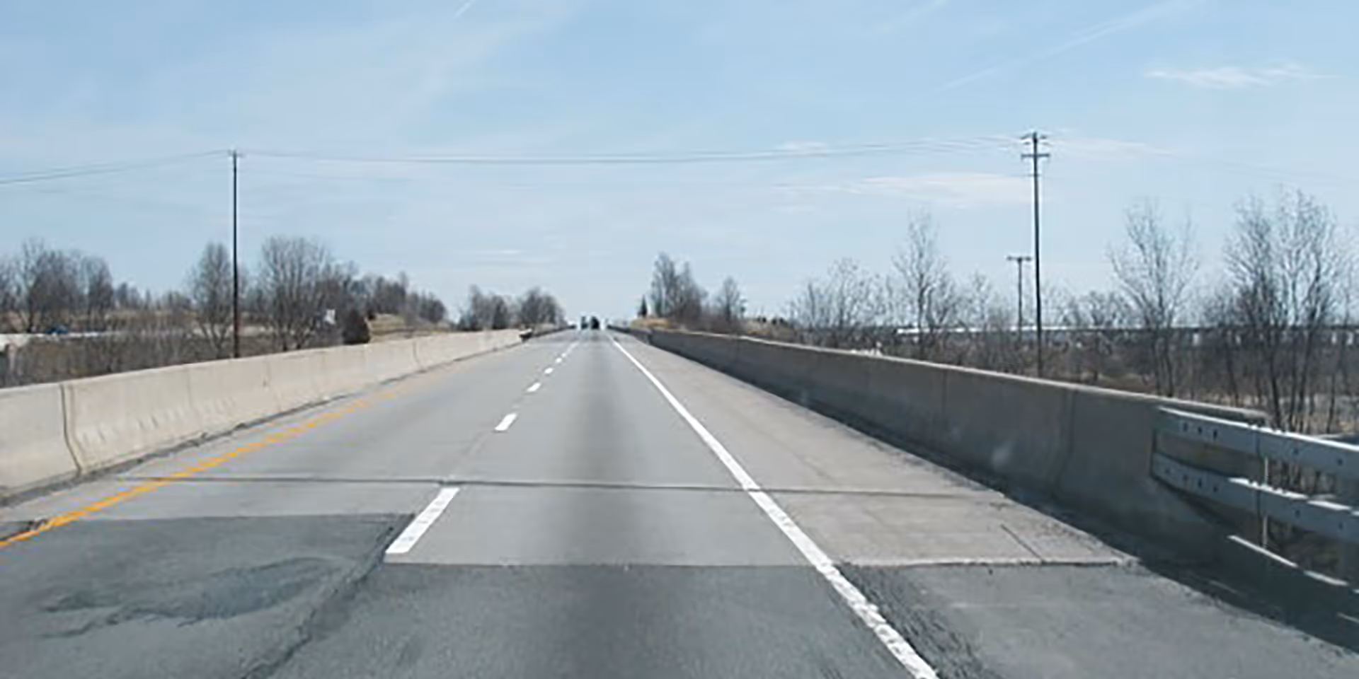 View of an empty, two-lane highway with concrete barriers on both sides and sparse trees in the distance.