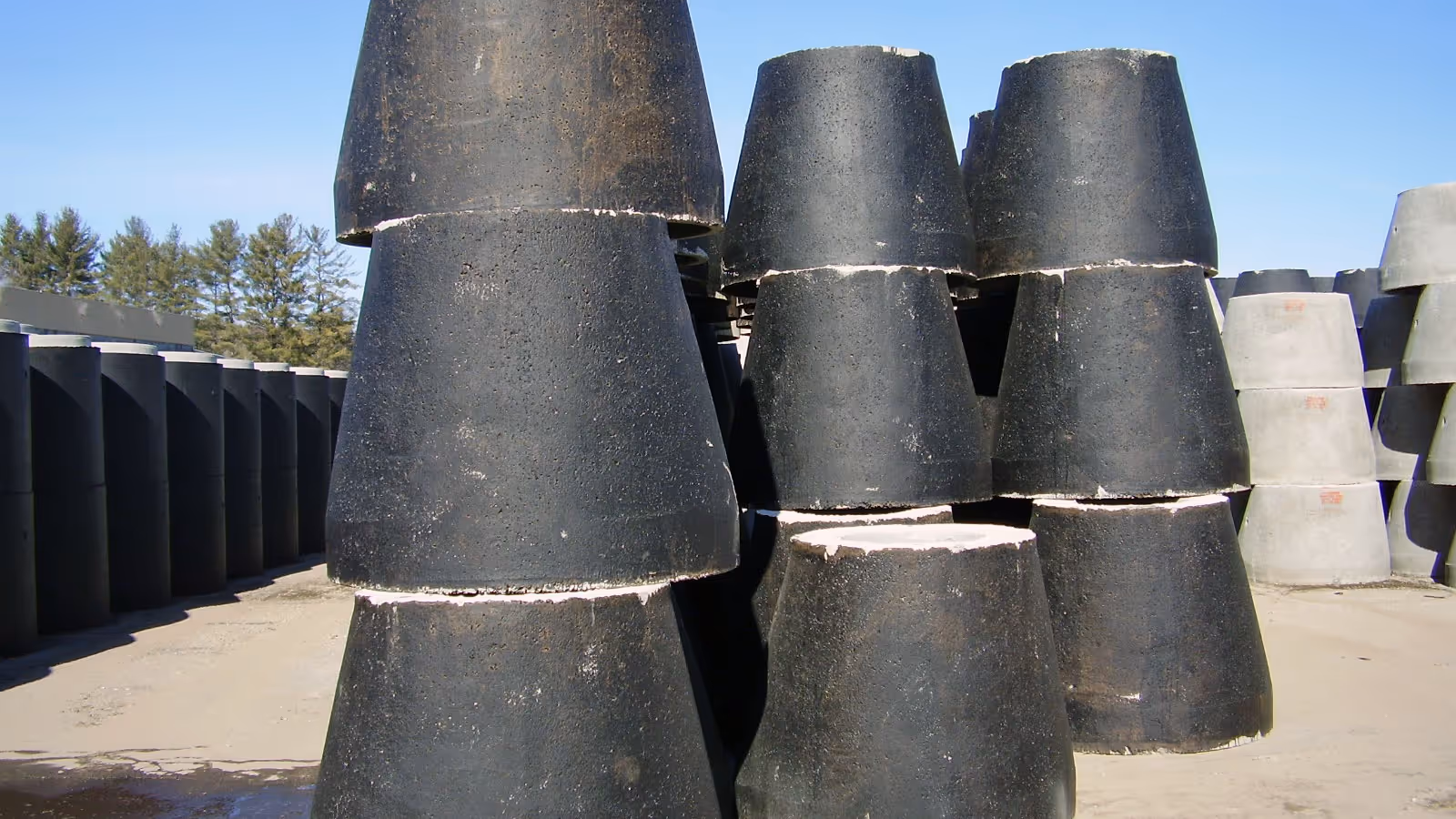Stacks of large, black, cylindrical concrete pipes are arranged outdoors on a sunny day.
