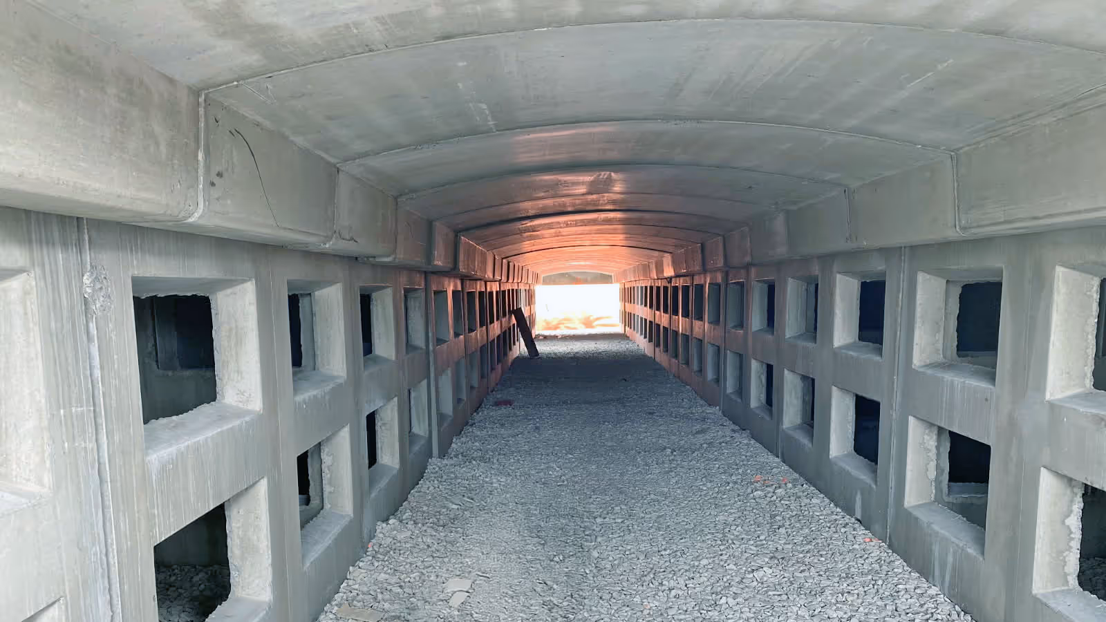 Inside view of a concrete tunnel with square openings on both sides and gravel covering the floor, light visible at the end.