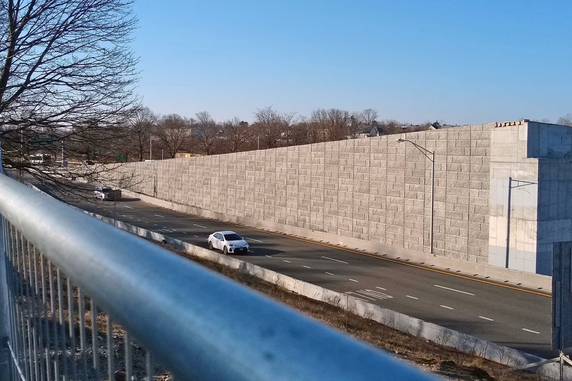 A white car drives down a nearly empty highway beside a tall concrete retaining wall on a clear day.