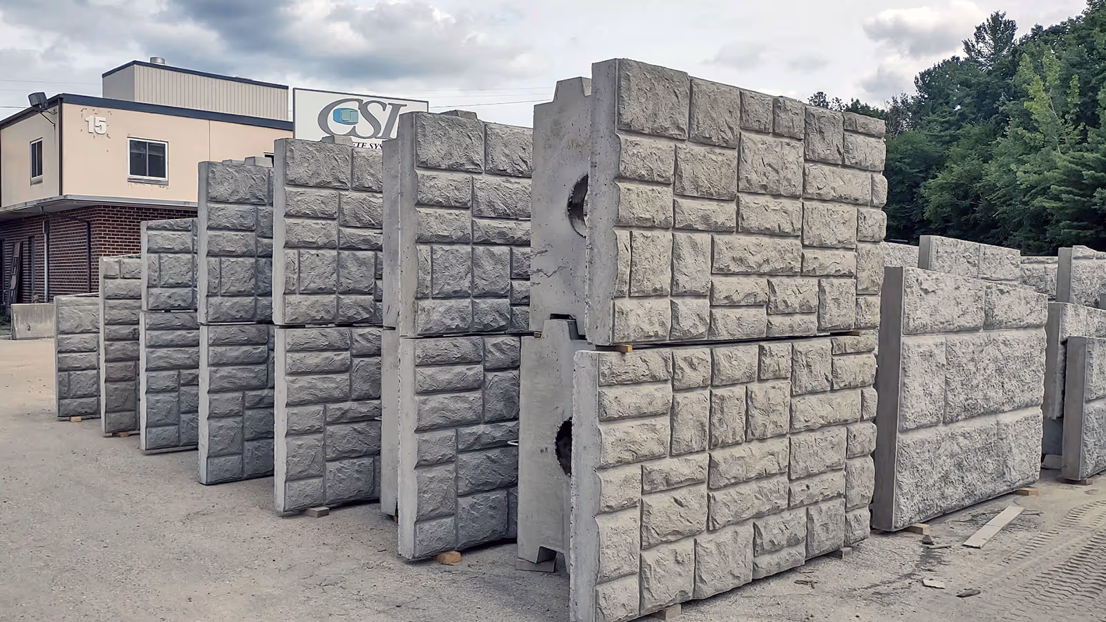 Large concrete panels with a textured stone pattern are lined up upright outdoors near a building and trees.