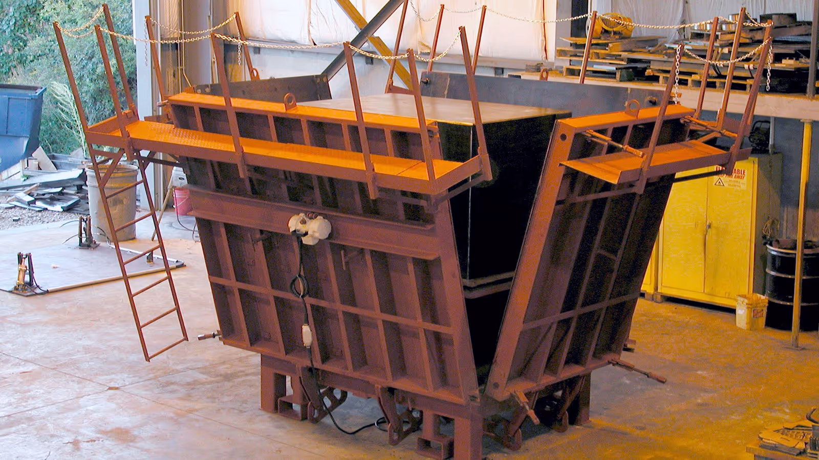 Large industrial steel mold with ladders and railings, set inside a warehouse with various equipment in the background.