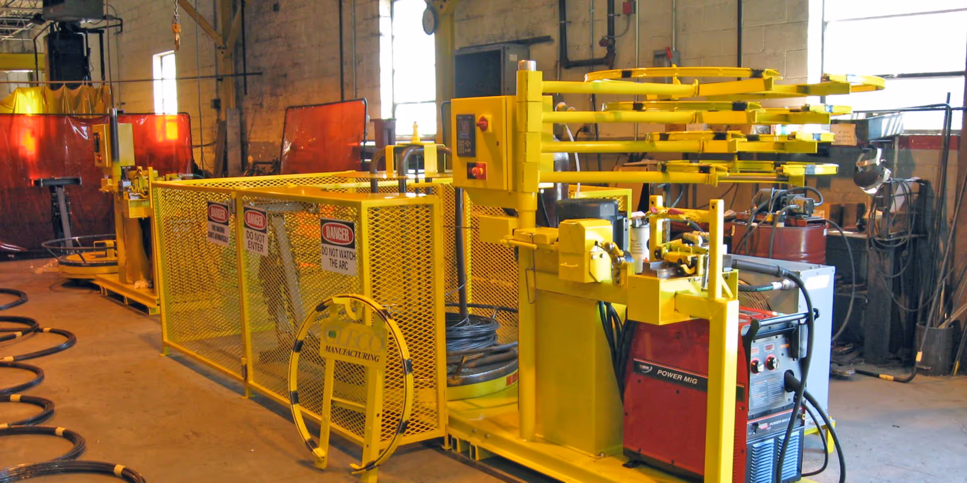 Yellow industrial bending machine and safety fencing in a workshop with various tools and equipment in the background.