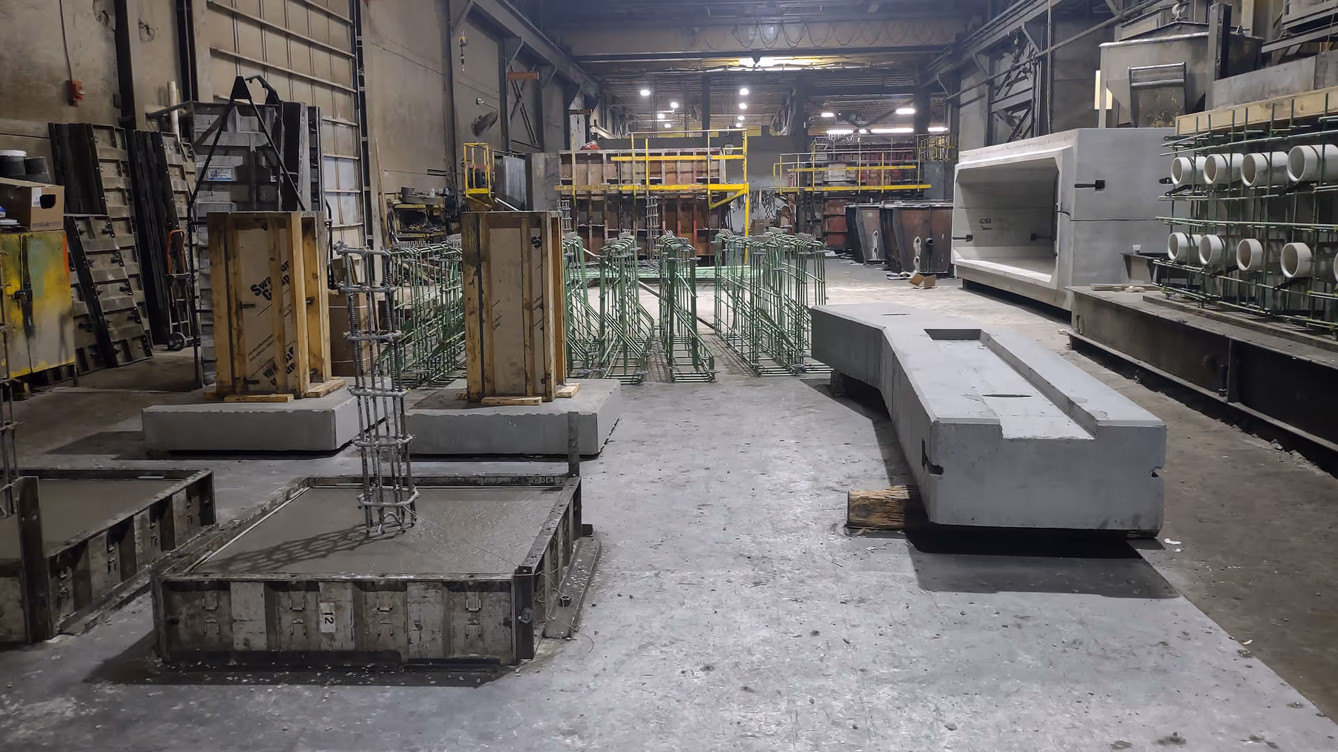 Industrial warehouse interior with concrete forms, metal reinforcement bars, and various construction materials.