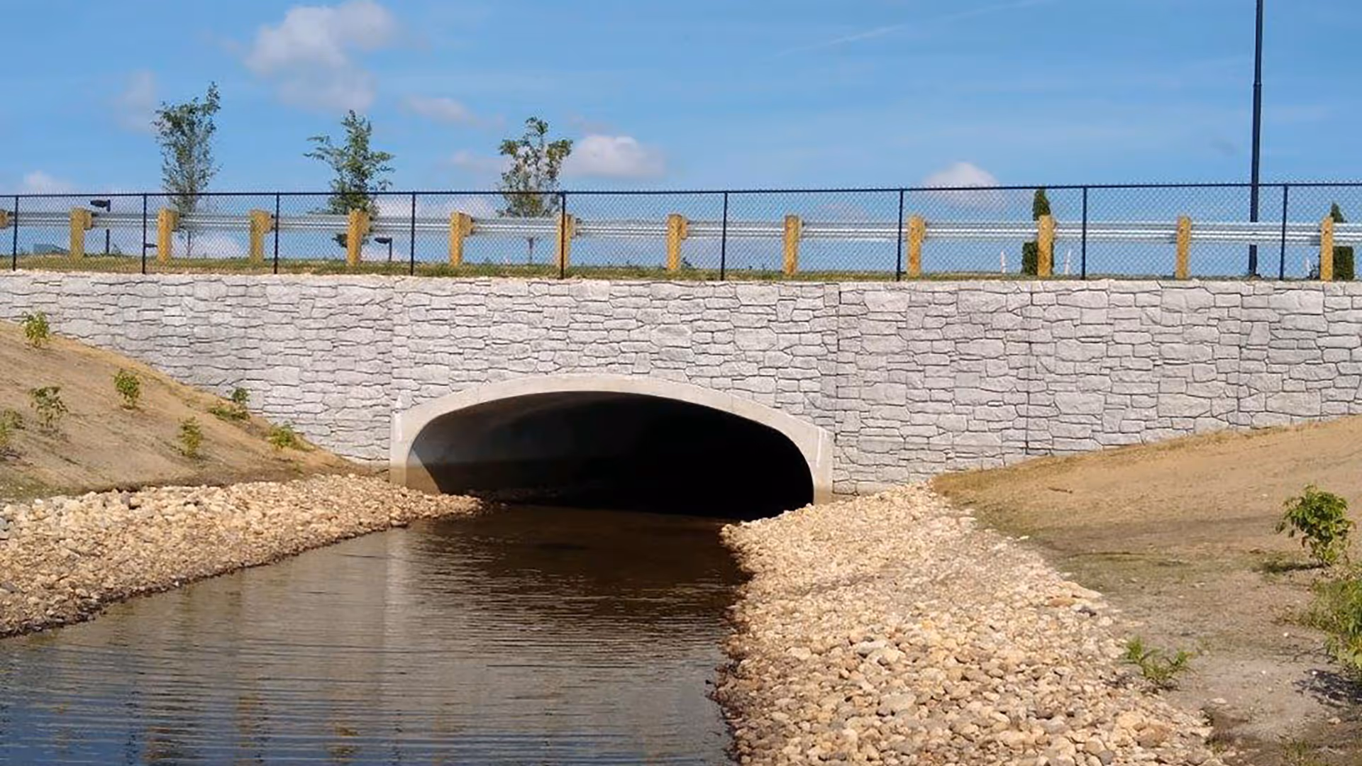 A concrete bridge with a stone facade crosses over a small creek lined with rocks and sparse vegetation.