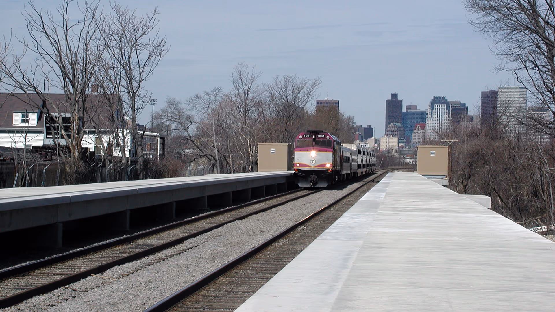 A commuter train approaches an empty platform with a city skyline visible in the background.
