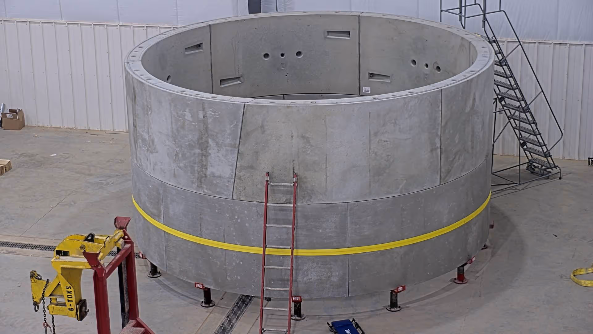 Large circular concrete structure with a yellow band sits indoors; ladders and equipment surround it on the floor.