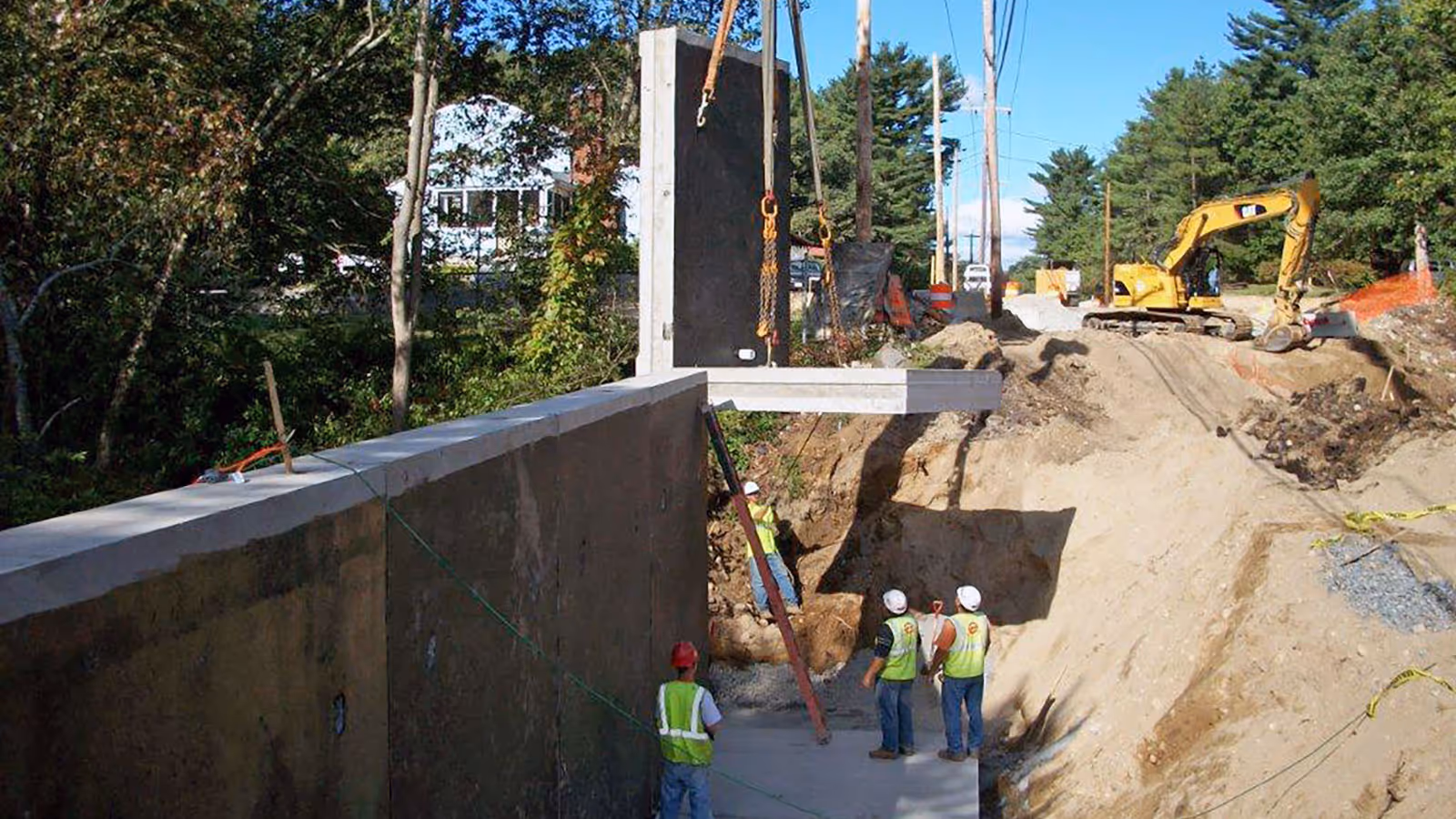 Construction workers in safety vests and helmets guiding the installation of a large precast concrete wall panel at an excavation site with a yellow excavator in the background.