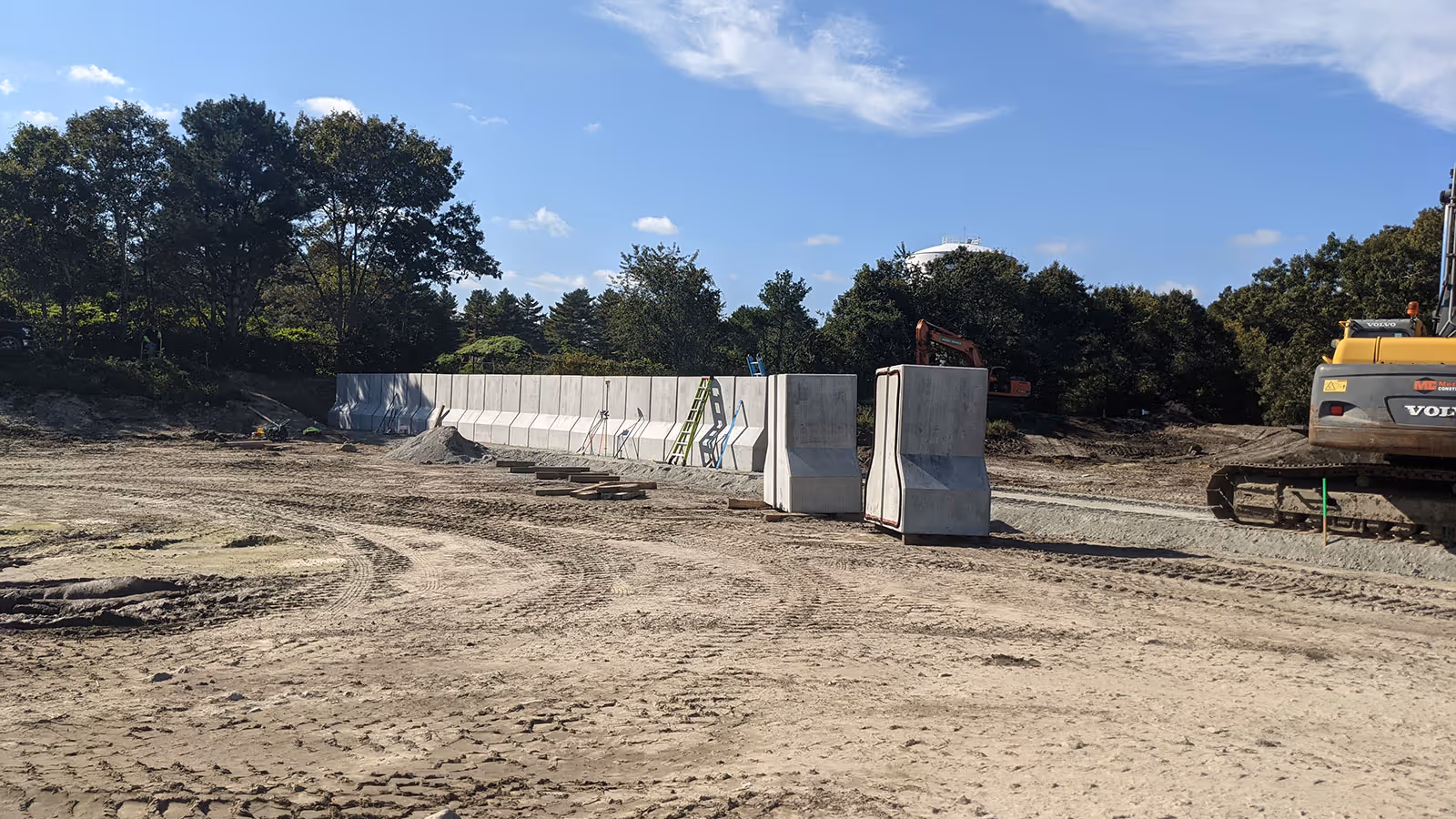 Construction site with stacked concrete barriers, machinery, and tire tracks on dirt ground under a partly cloudy sky.