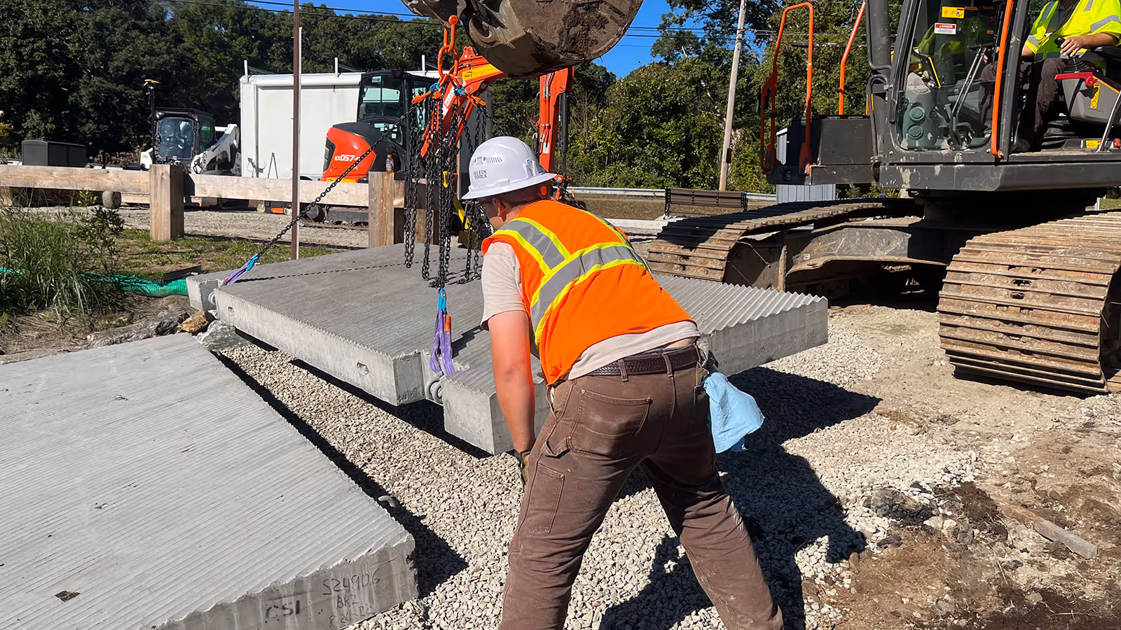 Construction worker in an orange safety vest and white helmet guiding a heavy concrete slab suspended by chains at a construction site.