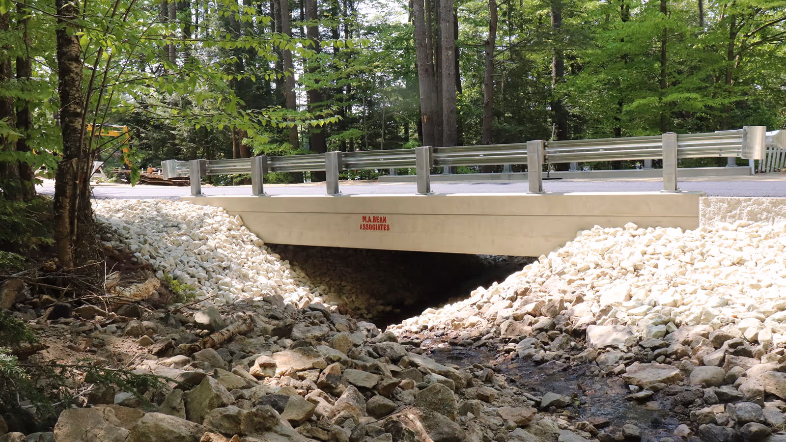 Small concrete bridge with metal guardrails over a rocky creek surrounded by green trees.