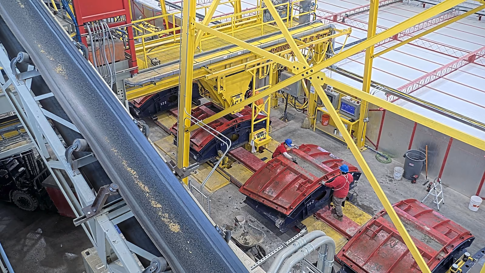 Two workers in red with blue helmets operating large red and yellow industrial machinery inside a factory.