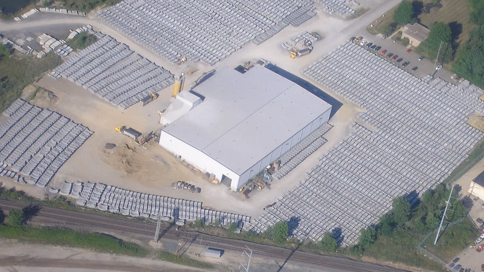 Aerial view of an industrial facility with a large warehouse surrounded by stacks of concrete pipes.