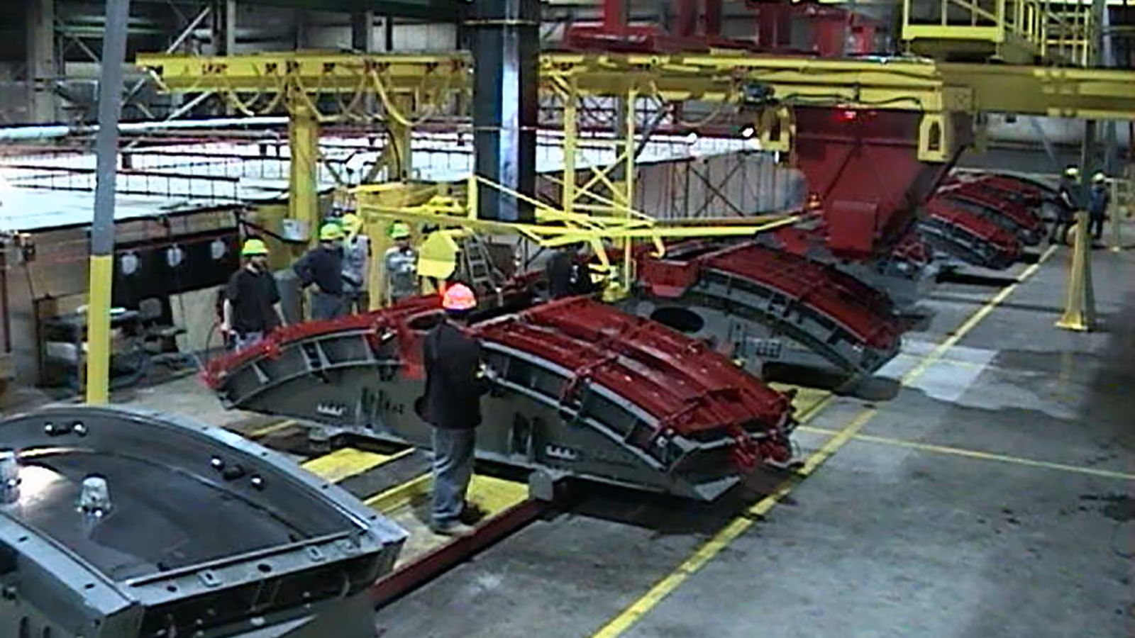 Worker inspecting large curved concrete tunnel segments lifted by yellow industrial crane inside factory.