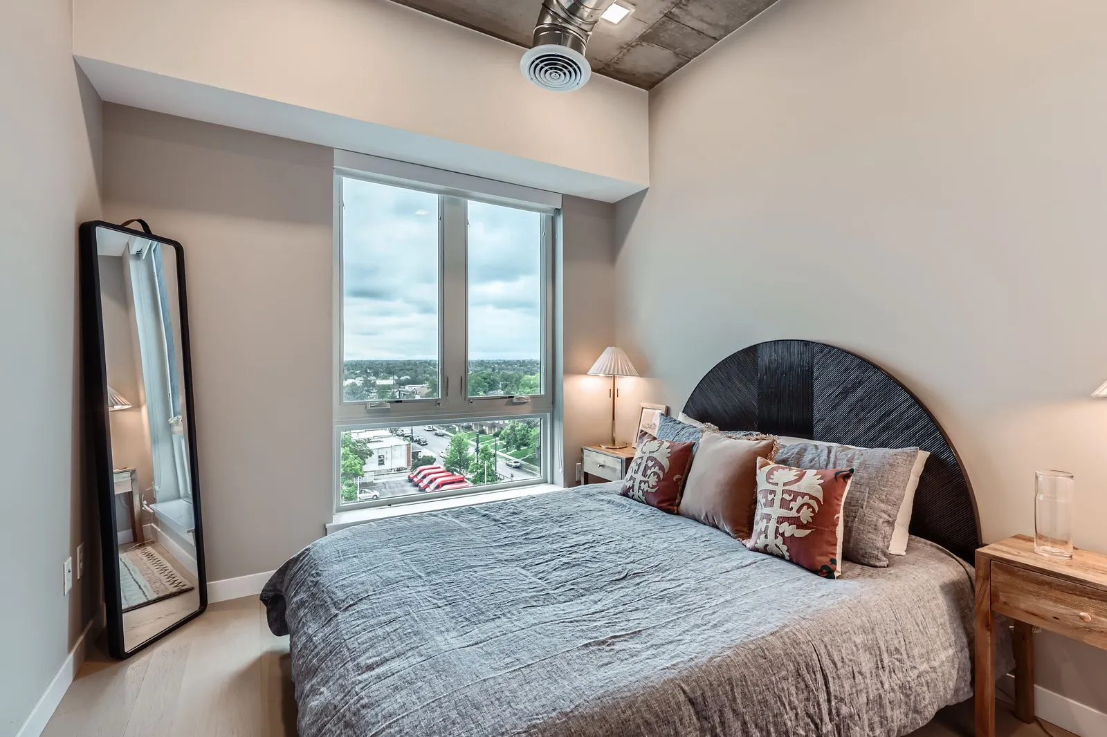 Modern bedroom with large window overlooking Sloans Lake and Denver cityscape, featuring a stylish bed and full-length mirror, at The Julian apartments.