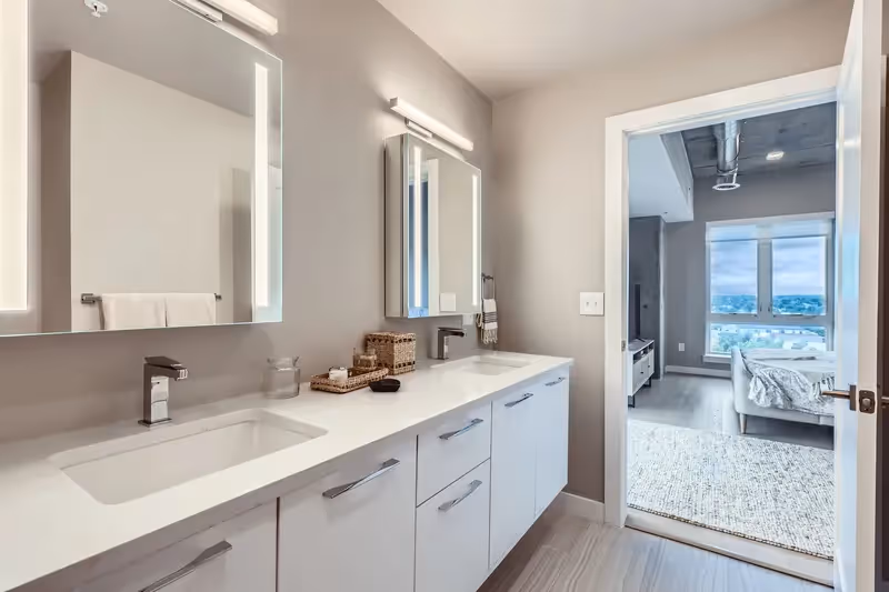 Modern bathroom in a luxury apartment at The Julian in Sloans Lake, Denver, featuring dual sinks, illuminated mirrors, and a view into a bedroom.