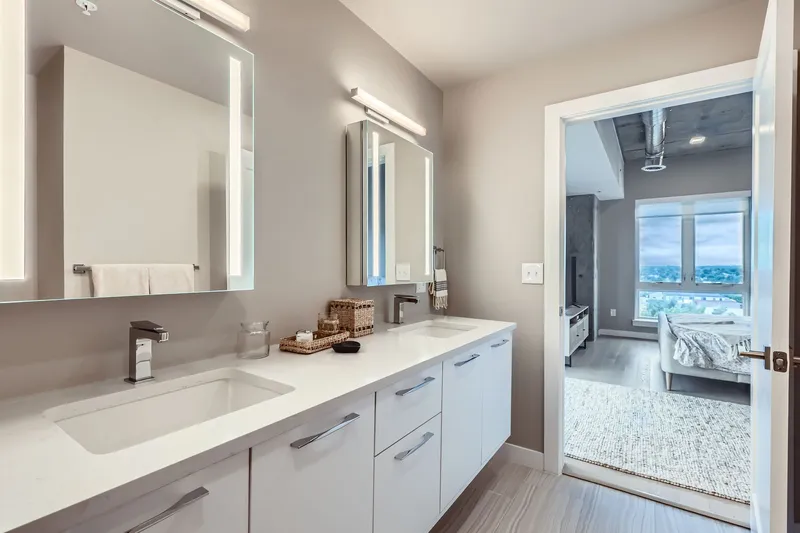 Modern bathroom in a luxury apartment at The Julian in Sloans Lake, Denver, featuring dual sinks, illuminated mirrors, and a view into a bedroom.