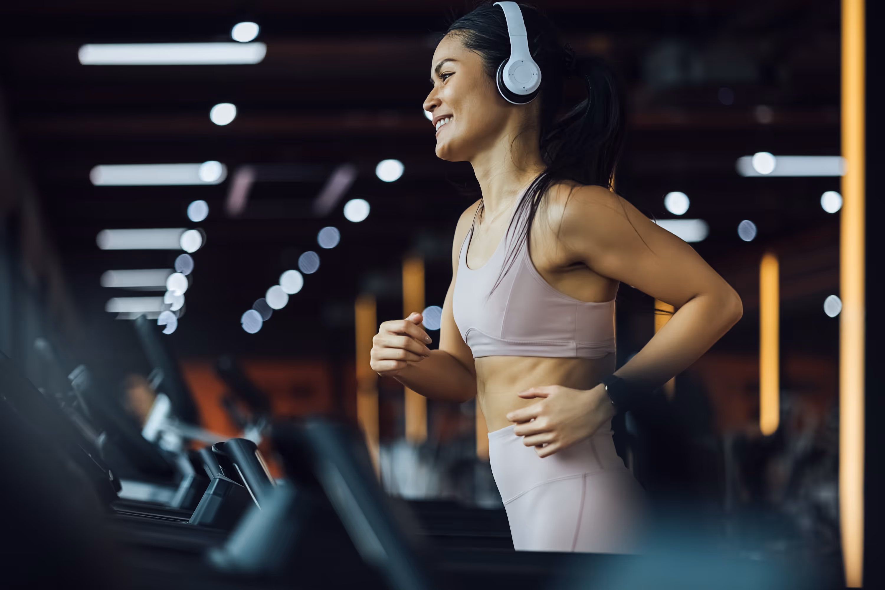 A smiling woman wearing headphones and running on a treadmill, representing the state-of-the-art fitness amenities at The Julian in Sloans Lake, CO.