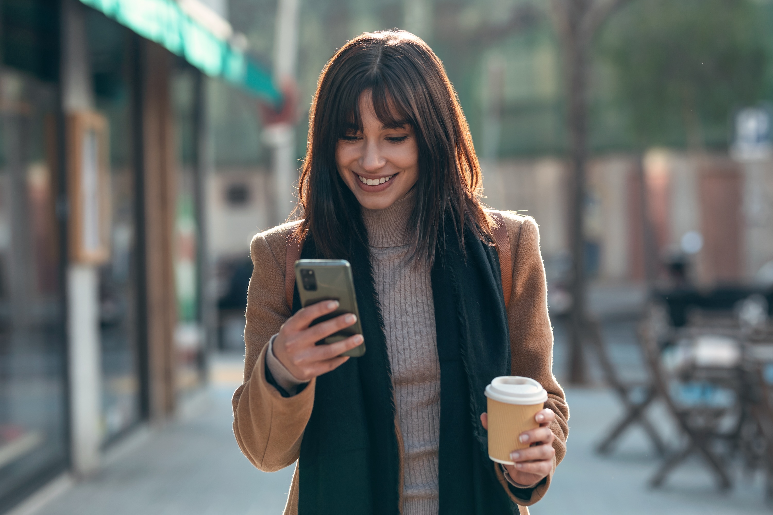A smiling woman in a coat and scarf, looking at her phone and holding a coffee cup while walking on a city street, representing the vibrant, walkable neighborhood of The Julian in Sloans Lake, CO.