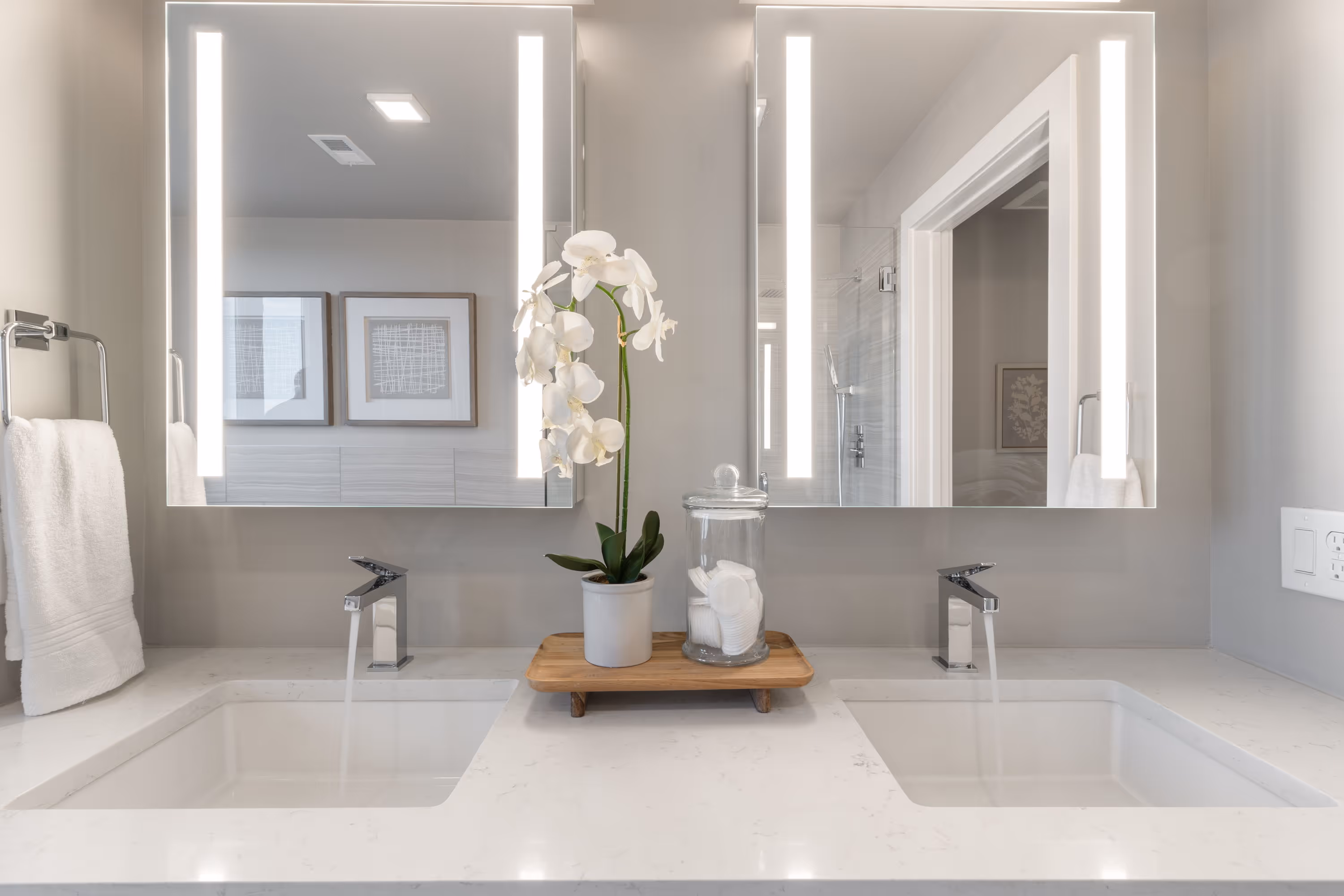 Modern bathroom in a luxury apartment at The Julian in Sloans Lake, Denver, featuring dual sinks, illuminated mirrors, and a view into a bedroom.