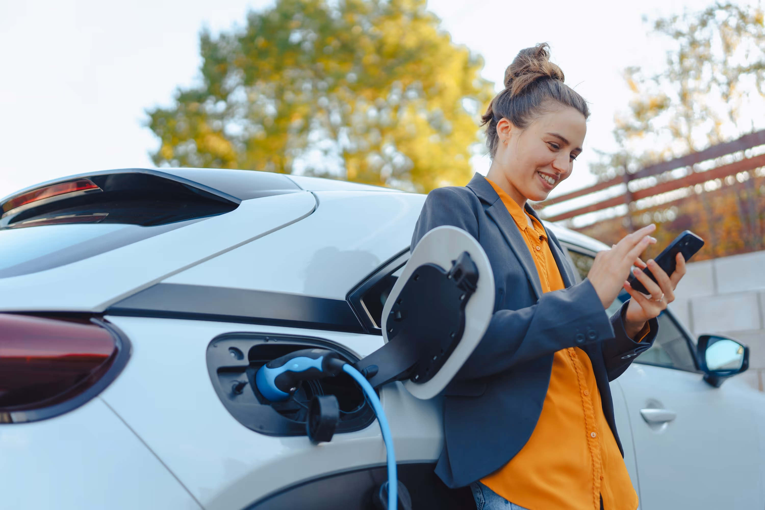 A smiling woman checks her phone while charging her electric vehicle (EV), representing the convenient EV charging stations and eco-friendly amenities at The Julian in Sloans Lake, CO.