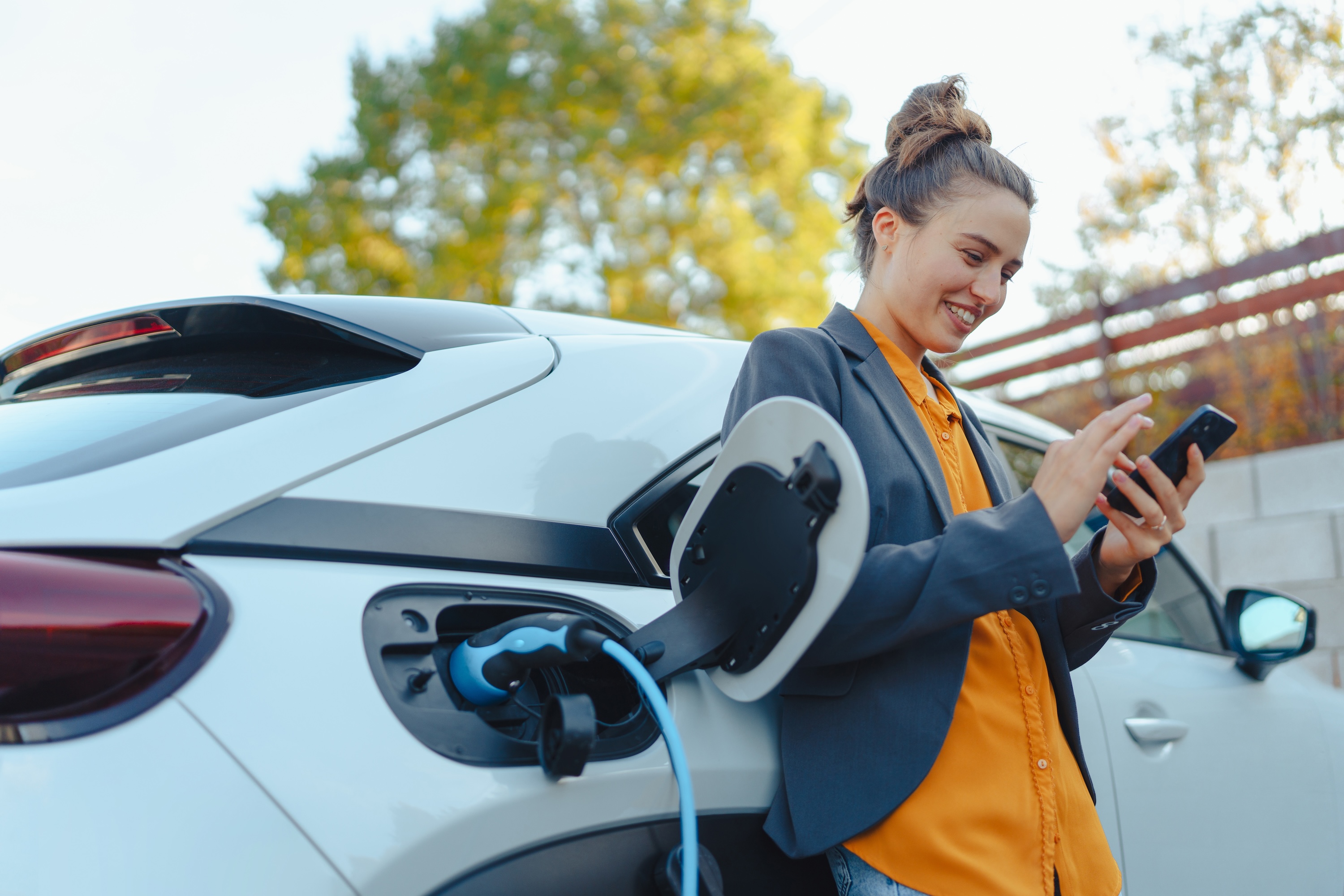 A smiling woman checks her phone while charging her electric vehicle (EV), representing the convenient EV charging stations and eco-friendly amenities at The Julian in Sloans Lake, CO.